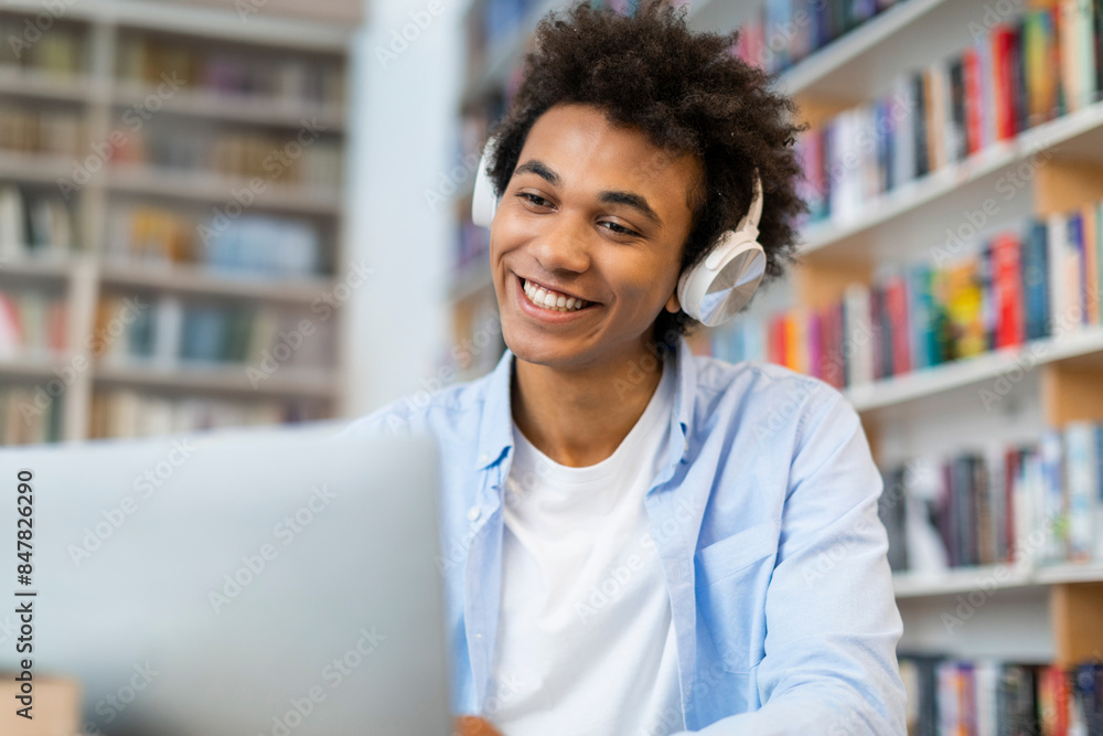 Cheerful Black student man in wireless headphones looking at laptop and smiling, talking on video call from college library Cheerful Black student man in wireless headphones looking at laptop and smiling, talking on video call from college library