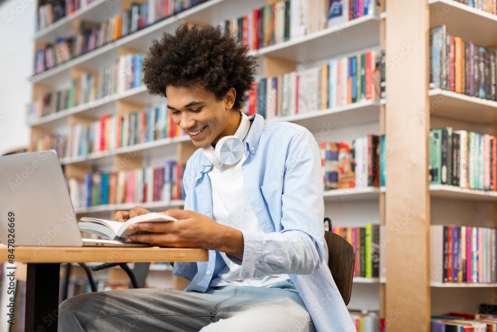 Positive Black male student reading book in university library sitting at desk with laptop, searching information for project Positive Black male student reading book in university library sitting at desk with laptop, searching information for project