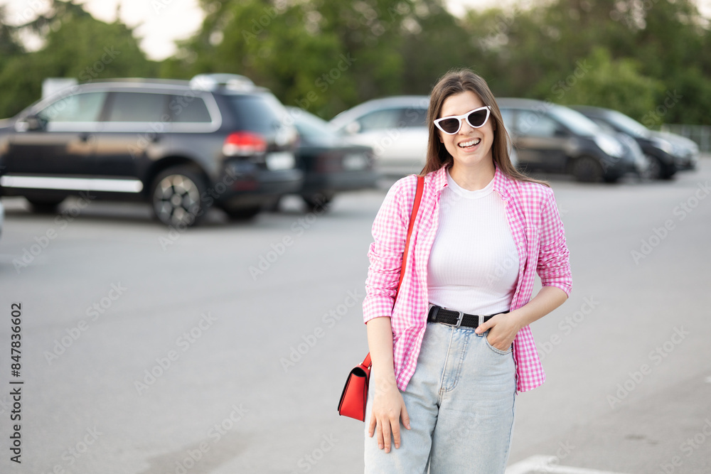 young woman wearing pink and white plaid shirt print vichy smiles while standing parking lot. white cat eyes sunglasses and red handbag casual outfit. fashionable stylish look of the day young woman wearing pink and white plaid shirt print vichy smiles while standing parking lot. white cat eyes sunglasses and red handbag casual outfit. fashionable stylish look of the day