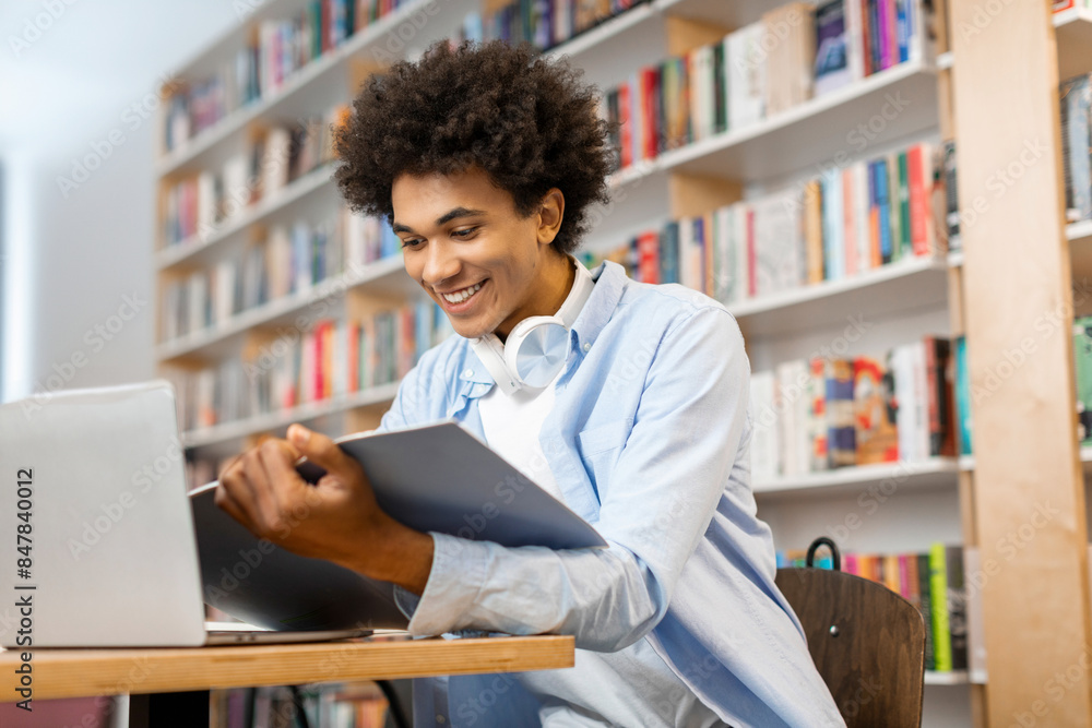 Latin student guy sitting in front of laptop and holding copybook, showcasing focused and traditional study approach with modern tools in library Latin student guy sitting in front of laptop and holding copybook, showcasing focused and traditional study approach with modern tools in library