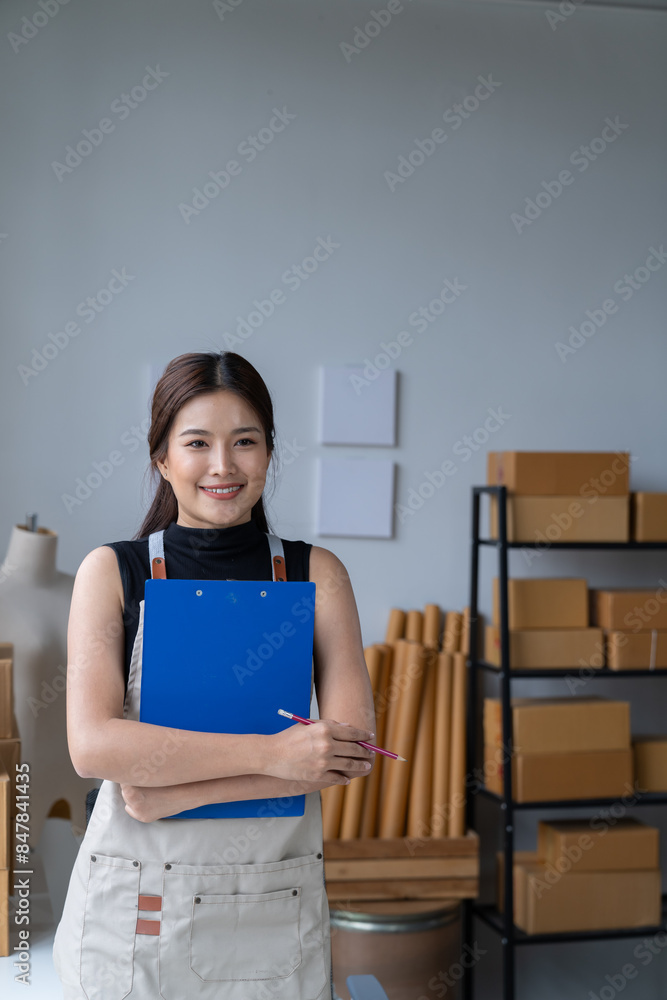 Obraz premium Delivery business. happy modern female in White Shirt checking order with laptop and holding a cup of coffee parcels using smartphone applications in the warehouse