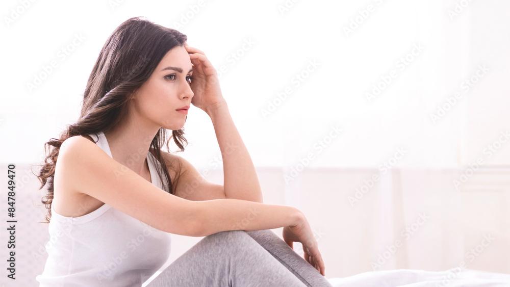 A woman with long brown hair sits on a bed, her hand resting on her forehead. Her expression is one of worry or sadness. She is wearing a white tank top and grey pants. A woman with long brown hair sits on a bed, her hand resting on her forehead. Her expression is one of worry or sadness. She is wearing a white tank top and grey pants.