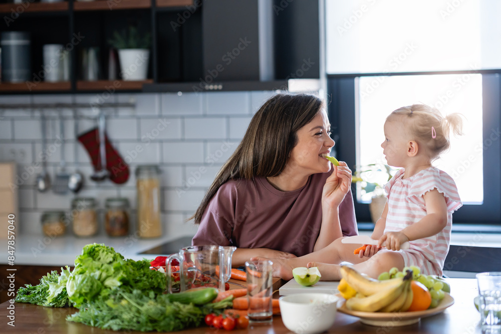 Cheerful mother and her cute little baby are eating healthy fruit and vegetables in the home kitchen Cheerful mother and her cute little baby are eating healthy fruit and vegetables in the home kitchen