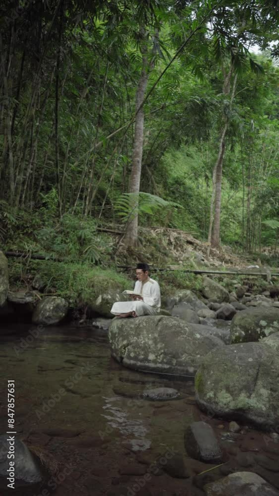 a young Asian Muslim who is reading the Koran on a rocky river, in a village near the forest during the month of Ramadan a young Asian Muslim who is reading the Koran on a rocky river, in a village near the forest during the month of Ramadan