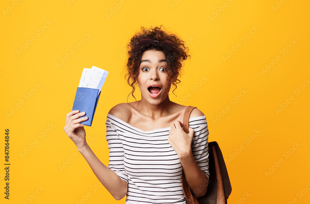 A joyful young African American woman with curly hair stands against a vivid yellow backdrop, holding what appears to be a pair of airline tickets and a passport A joyful young African American woman with curly hair stands against a vivid yellow backdrop, holding what appears to be a pair of airline tickets and a passport