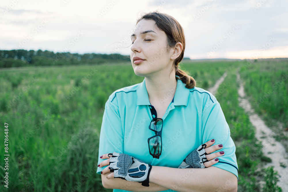 Woman in a blue shirt standing in a field with her arms crossed. She is wearing cycling gloves and has sunglasses hanging from her shirt. The woman appears to be resting after a bike ride.