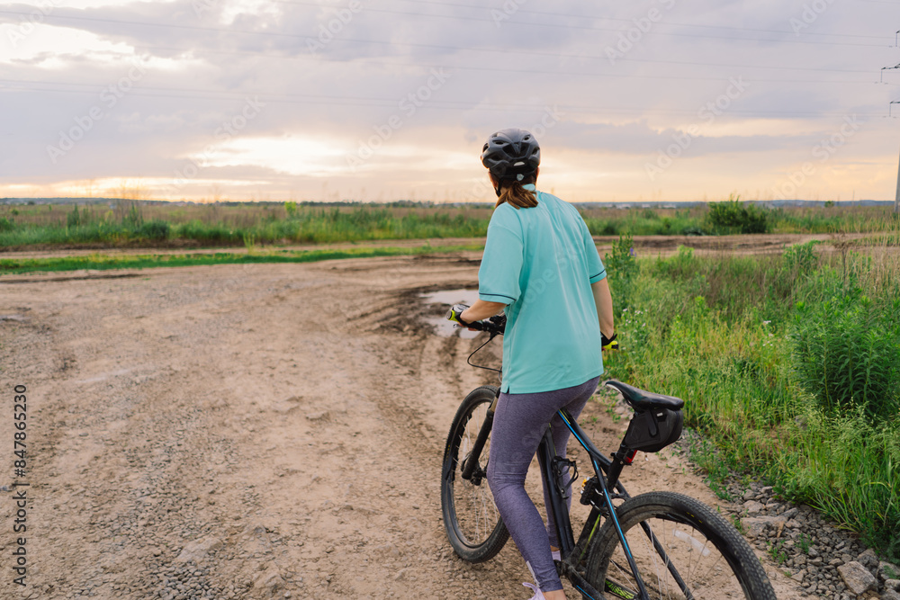 Fototapeta premium A woman wearing a helmet rides a black mountain bike along a dirt path. The path runs through an open grassy field, with houses and trees visible in the background.