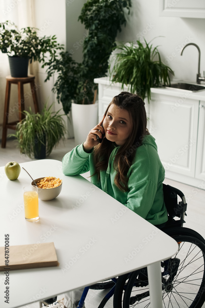 A young brunette woman with a disability sits in a wheelchair at her kitchen table, talking on her cell phone. A young brunette woman with a disability sits in a wheelchair at her kitchen table, talking on her cell phone.