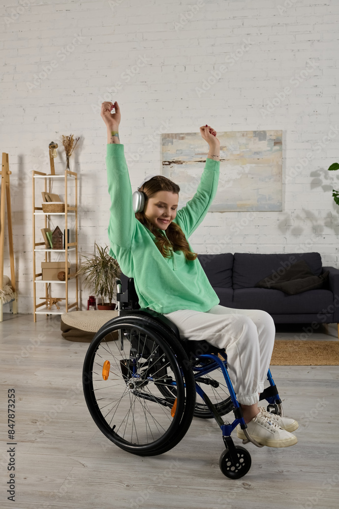 Obraz premium A young brunette woman with a disability sits in a wheelchair in her living room and dances along to music playing in her headphones.