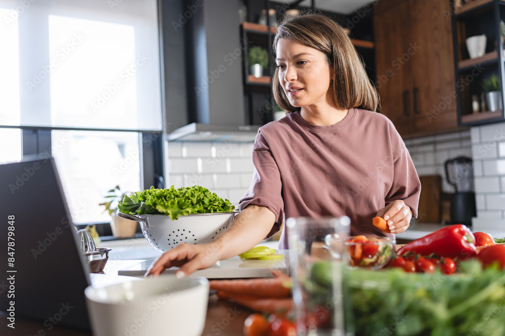A young, pretty, cheerful woman is looking at a laptop computer while preparing food at home, following a cooking recipe online A young, pretty, cheerful woman is looking at a laptop computer while preparing food at home, following a cooking recipe online