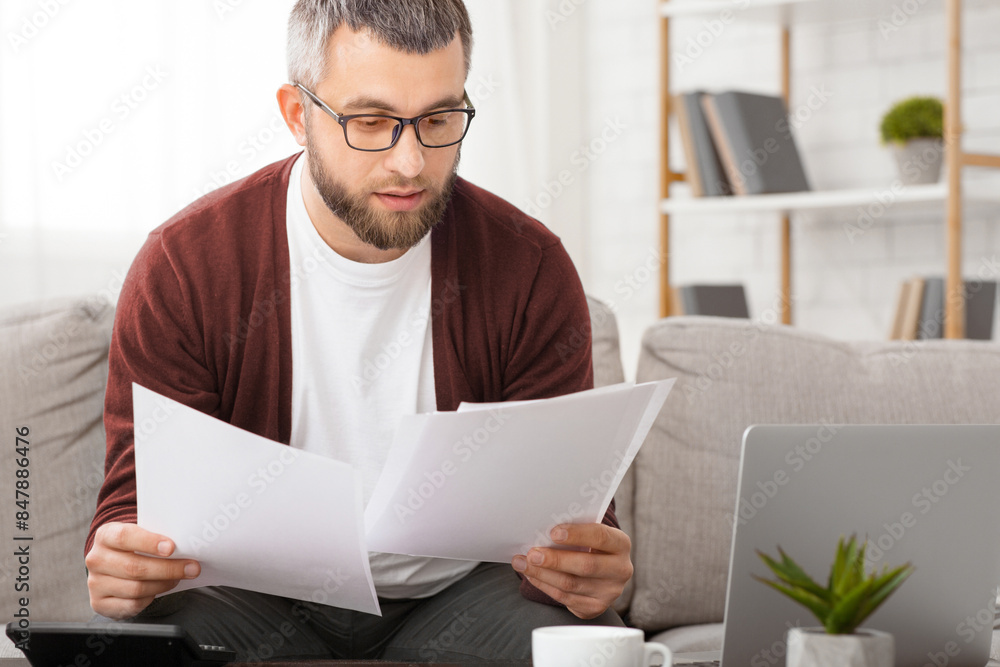 Concentrated man is seated on a couch, focused on reading a piece of paper in his hand. He appears concentrated and engaged in the content of the document. Concentrated man is seated on a couch, focused on reading a piece of paper in his hand. He appears concentrated and engaged in the content of the document.