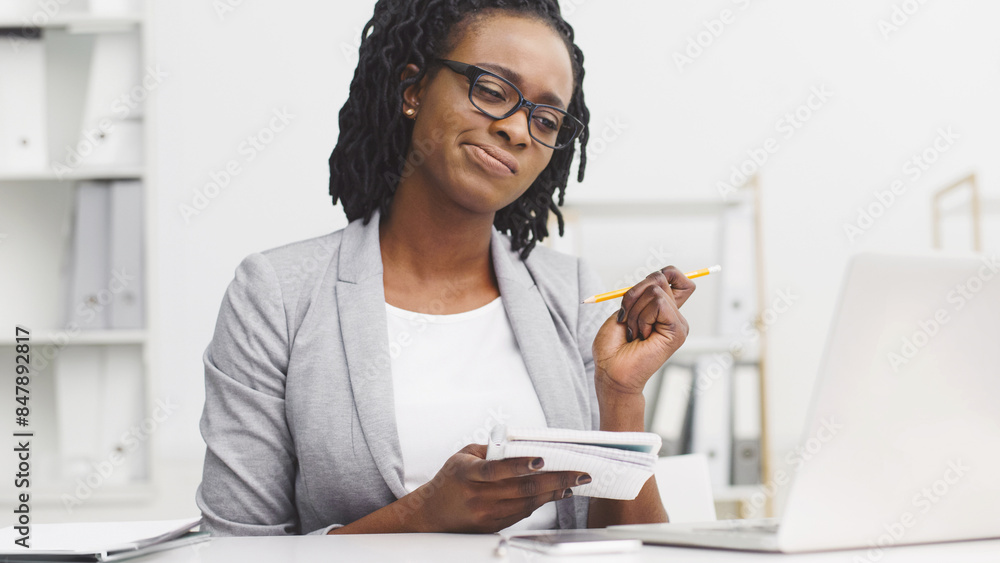 Thoughtful African American lady working on report, sitting at desk, writing in notepad, looking at laptop screen at modern office Thoughtful African American lady working on report, sitting at desk, writing in notepad, looking at laptop screen at modern office