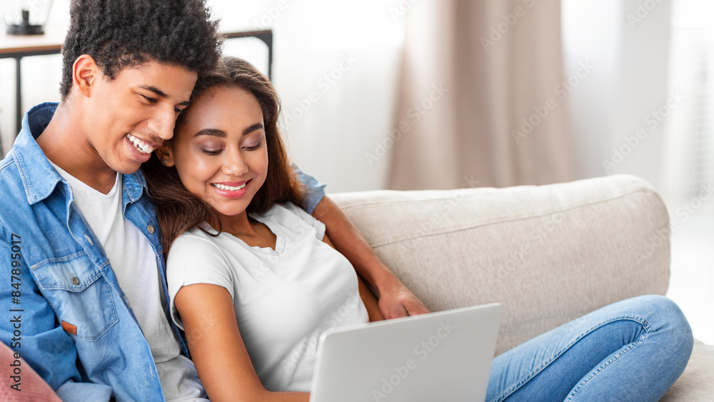 African American teen guy and girl are relaxing on a couch in a living room. The guy is sitting with his arm around the girl, who is looking at a laptop in her lap. They appear happy and comfortable. African American teen guy and girl are relaxing on a couch in a living room. The guy is sitting with his arm around the girl, who is looking at a laptop in her lap. They appear happy and comfortable.
