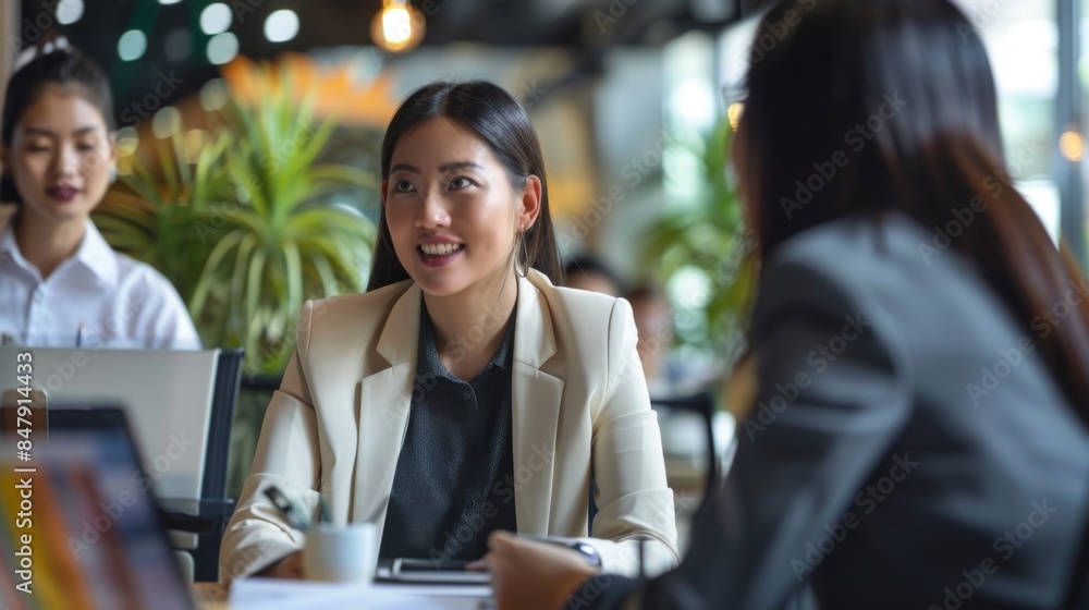 Fototapeta premium A young woman in a white blazer sits at a table in a cafe, smiling while listening to another woman
