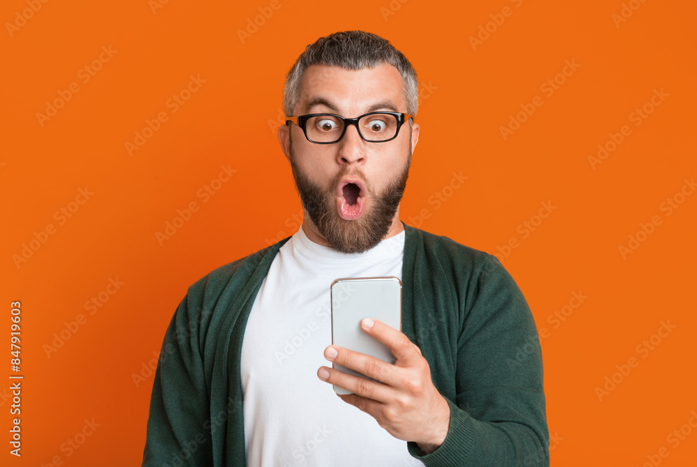 A bearded man with glasses is expressing shock as he looks at his smartphone. He is dressed casually in a green sweater and a white t-shirt. The background is a solid orange color A bearded man with glasses is expressing shock as he looks at his smartphone. He is dressed casually in a green sweater and a white t-shirt. The background is a solid orange color