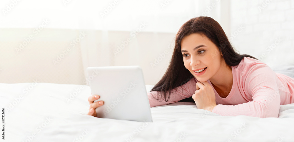 A woman is lying comfortably on a white bed, smiling as she uses a tablet device. The cozy bedroom features soft natural light coming through the window, creating a peaceful and inviting atmosphere. A woman is lying comfortably on a white bed, smiling as she uses a tablet device. The cozy bedroom features soft natural light coming through the window, creating a peaceful and inviting atmosphere.