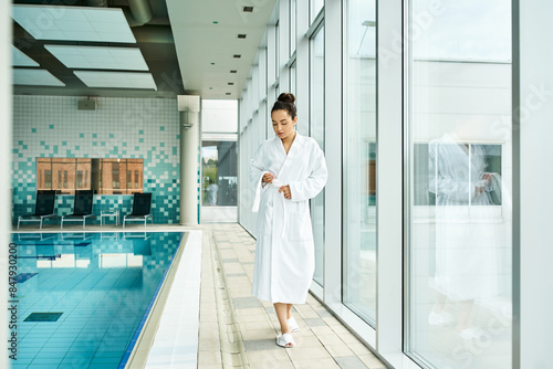 A young, beautiful brunette woman in a bathrobe standing beside an indoor swimming pool. © LIGHTFIELD STUDIOS