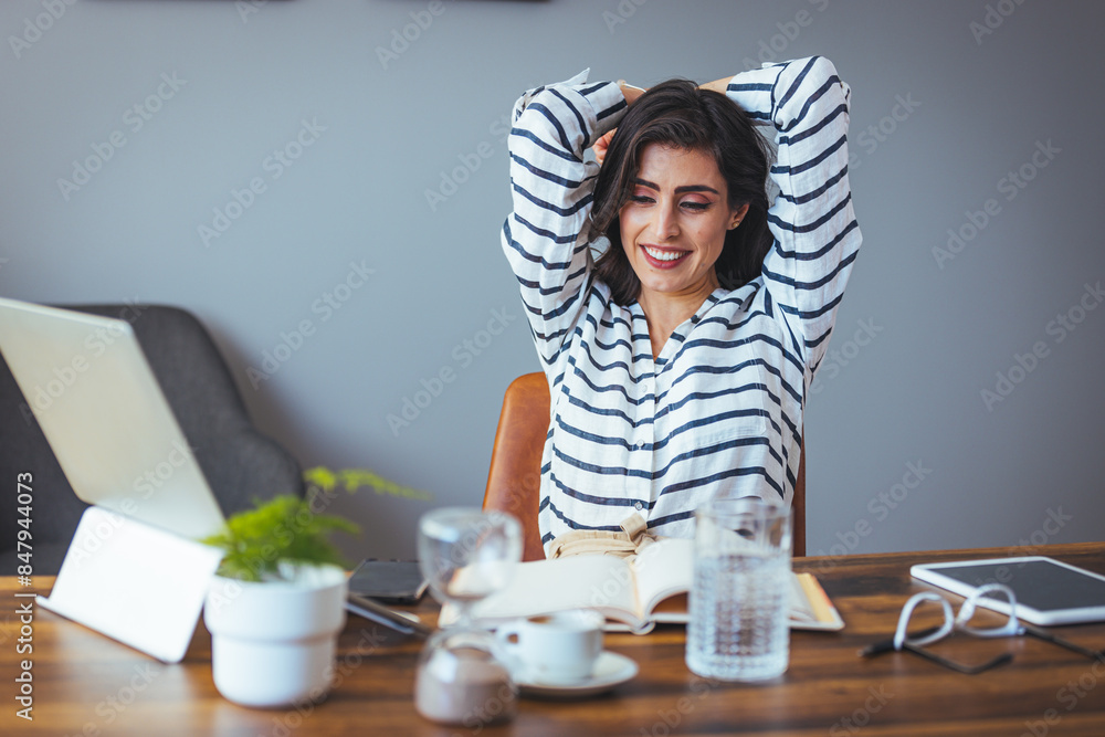 A smiling Caucasian woman in striped business attire stretches for relaxation amidst her busy office environment, surrounded by work essentials like a laptop and notepad. A smiling Caucasian woman in striped business attire stretches for relaxation amidst her busy office environment, surrounded by work essentials like a laptop and notepad.