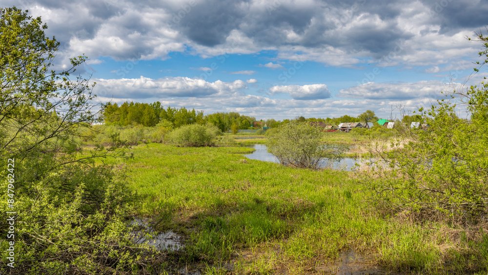 A tranquil spring scene with lush green meadows, a winding creek, and a glimpse of homes in the distance. The sky is filled with fluffy white clouds against a backdrop of bright blue. A tranquil spring scene with lush green meadows, a winding creek, and a glimpse of homes in the distance. The sky is filled with fluffy white clouds against a backdrop of bright blue.