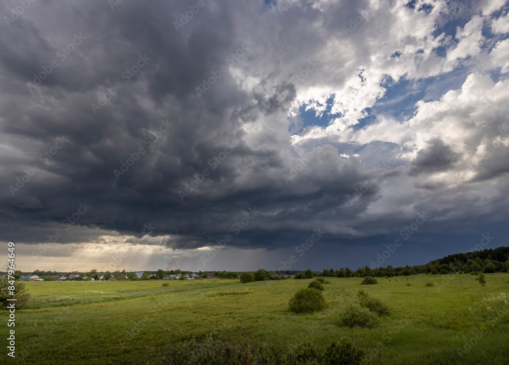 A vast, ominous storm cloud looms over a verdant meadow, casting dramatic shadows across the landscape.The sun breaks through the cloud in places, illuminating the scene with a fleeting burst of light