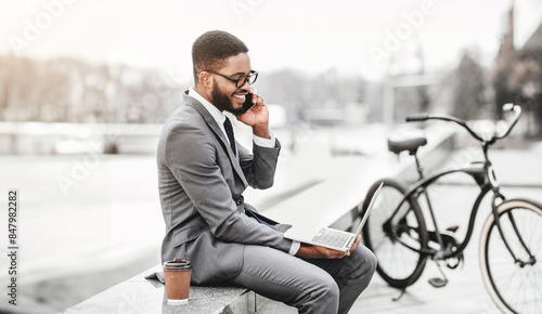 An African American businessman is sitting outdoors with his laptop, talking on his phone, and his bicycle next to him. The mood is productive and relaxed, blending work with nature.