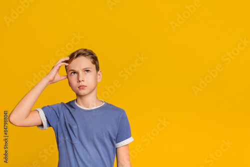 Pensive teenage boy looking up with thoughtful expression, holding hand on head, trying to remember something, orange background with free space photo