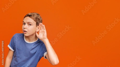 Curious snoopy schoolboy holding hand at his ear, trying to overhear private conversation, posing at orange studio background photo