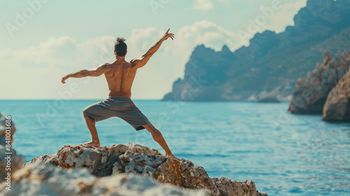 Shirtless man practicing yoga on rocky seashore with mountains in background photo