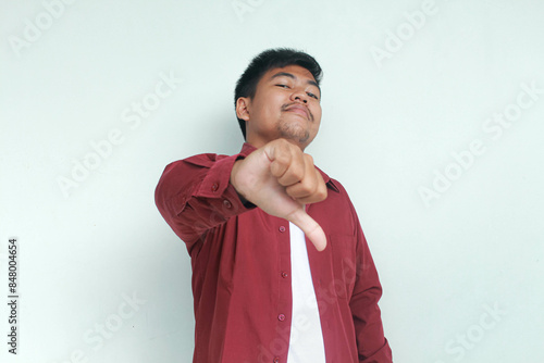 Proud Asian man wearing red shirt gives thumbs down hand gesture of don't agree, isolated by grey background.