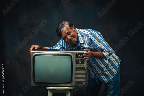 An elderly man hugging an old TV set on a chair on black background in the studio.