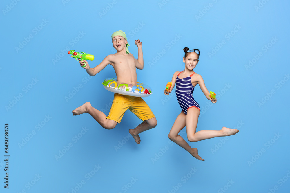 Obraz premium Happy brother and sister in swimwear jumping with water guns on blue studio background