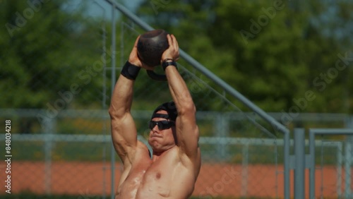 Young man is training on sports field and doing kettlebell press with both hands. Strength exercises help in developing beautiful muscular body. Slow motion