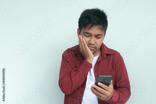 Portrait of unhappy and stressed Asian man in red shirt holding mobile phone with sad expression on face while scratching forehead. Isolated image on gray background.