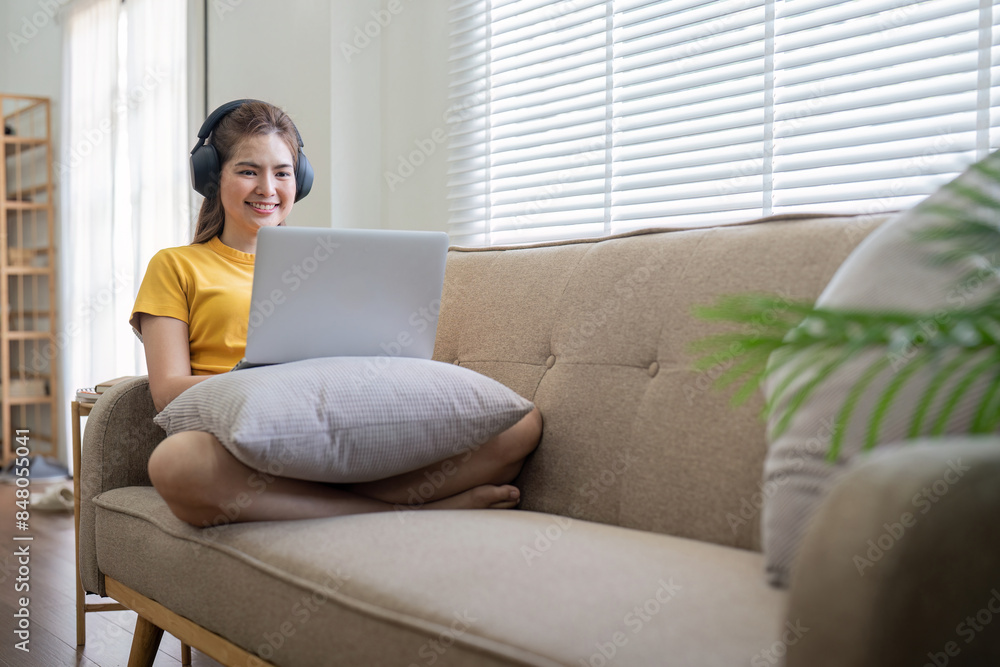 Young Woman Enjoying Music at Home with Headphones While Using Laptop on Sofa in Bright Modern Living Room Young Woman Enjoying Music at Home with Headphones While Using Laptop on Sofa in Bright Modern Living Room