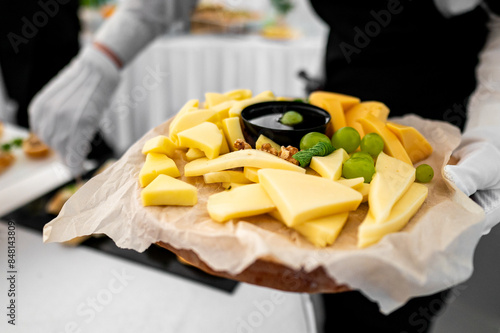Close-up of a cheese platter with various cheeses, grapes, and nuts on a wooden board. Served by a person in white gloves against a blurred background photo