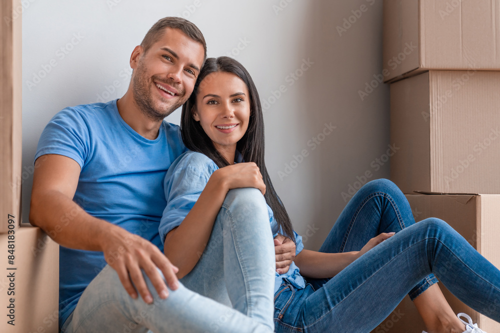 Fototapeta premium Happy family of two sitting on floor near wall around boxes after buying house. Moving Day. Lovely couple wife and husband sitting surrounded with boxes on the floor in new home