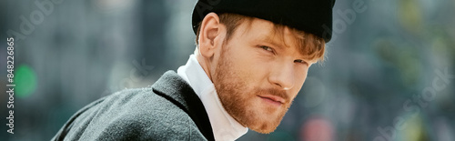 A young red-haired man with a beard, in debonair attire, strolling through the city wearing a black hat.