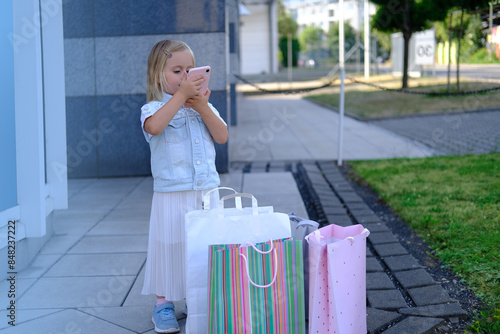 happy child of 3-4 years old, girl in fashionable clothes standing with craft bags full purchases on street, surfing on phone, happy shopping on Black Friday, young shopaholic, little fashion-girl photo