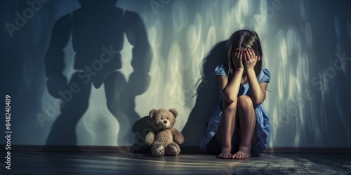 Stop violence and abused children. A sad little girl sits on the floor in a dark room, covering her head with her hands and a stuffed toy next to her. The concept of human trafficking.