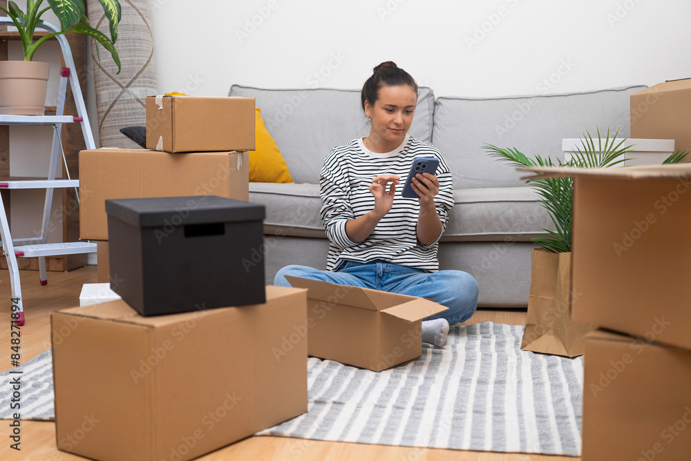 Woman scrolls social media in smartphone with carton boxes around near sofa in house waits for husband from work to unpack things to decorate contemporary apartment Woman scrolls social media in smartphone with carton boxes around near sofa in house waits for husband from work to unpack things to decorate contemporary apartment
