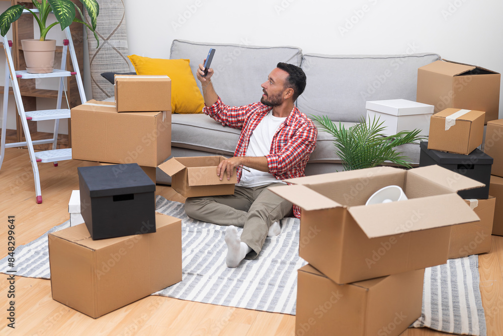 A youthful man unpacks moving boxes, shares his new home via video call, captures a selfie, embracing the essence of relocation. A youthful man unpacks moving boxes, shares his new home via video call, captures a selfie, embracing the essence of relocation.