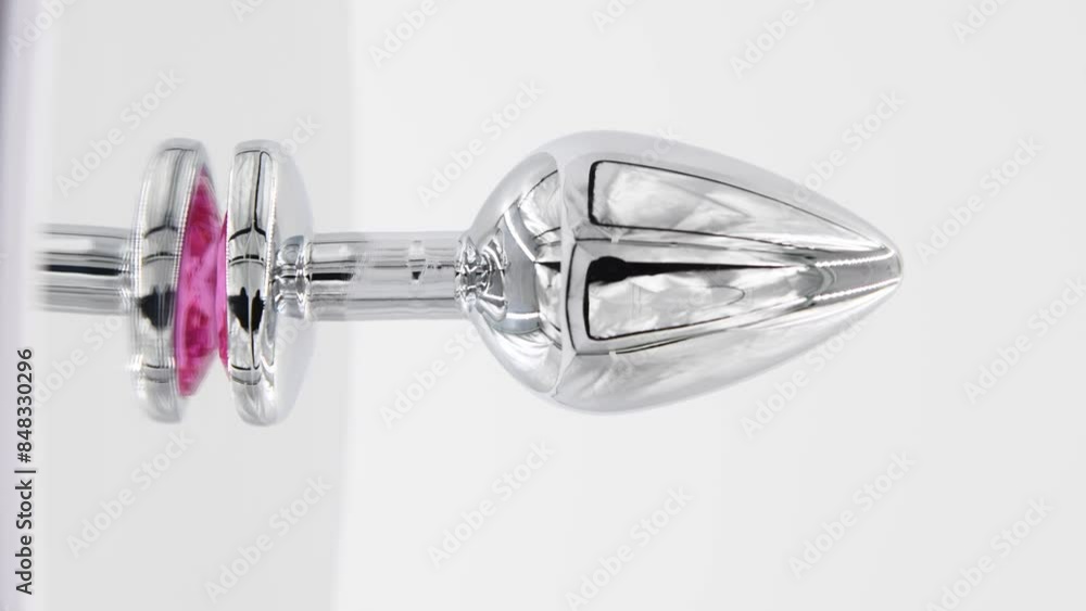 Close-up side view of metallic butt plug with pink heart shaped crystal spinning on mirror surface against white background. Soft focus. Real time vertical video. Adult sex toys theme. Close-up side view of metallic butt plug with pink heart shaped crystal spinning on mirror surface against white background. Soft focus. Real time vertical video. Adult sex toys theme.