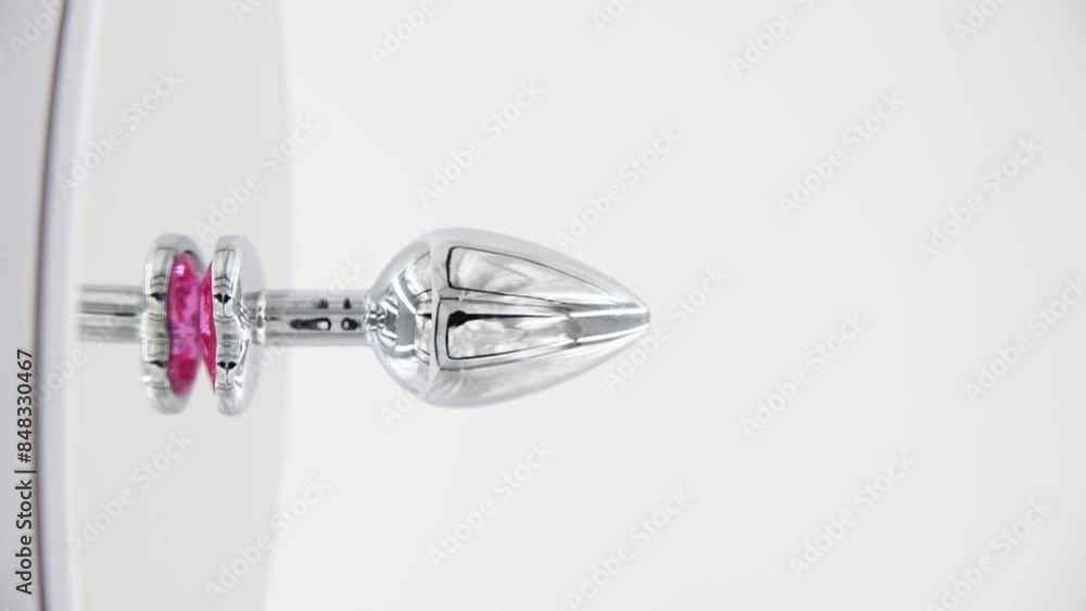 Close-up side view of metallic butt plug with pink heart shaped crystal spinning on mirror surface against white background. Soft focus. Real time vertical video. Adult sex toys theme. Close-up side view of metallic butt plug with pink heart shaped crystal spinning on mirror surface against white background. Soft focus. Real time vertical video. Adult sex toys theme.