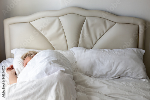 blonde mature man sleeps on a bed with a white headboard under a white blanket in the bedroom. Comfortable sleep.