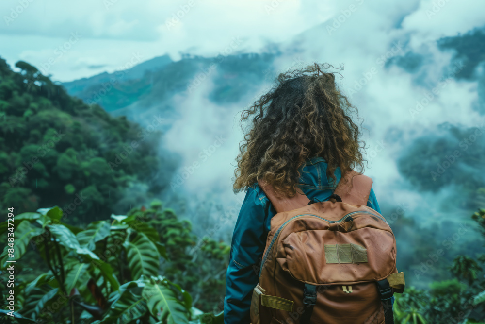 Naklejka premium Back view of a woman with curly hair and backpack hiking through a misty mountain forest, surrounded by lush greenery. Female traveler enjoying nature landscape