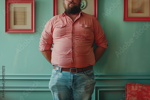 Plump, bearded man in jeans with pockets full of items that make his jeans pockets stick out. Phone and keys stick out from pants pockets of middle aged man posing in pink shirt against wall