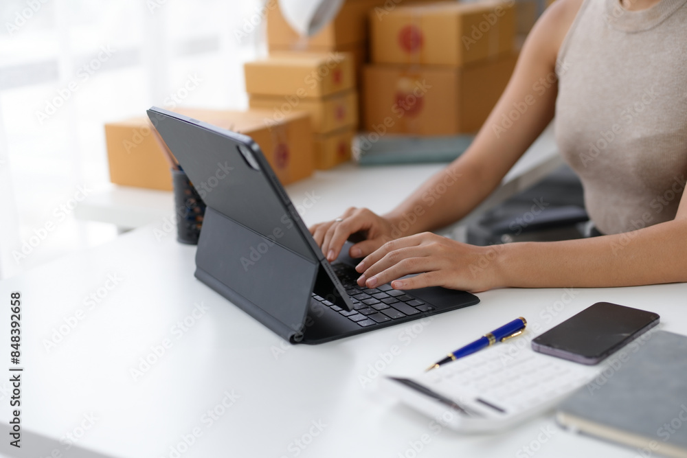 Fototapeta premium Woman working on a tablet at a desk with shipping boxes in the background, illustrating e-commerce and small business operations.