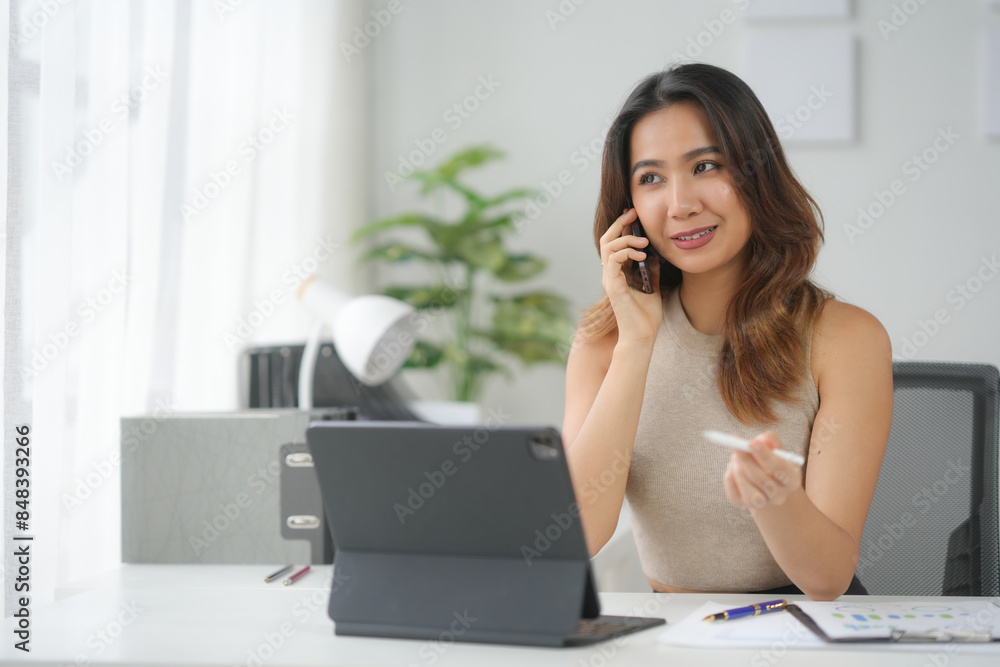 Young professional woman talking on phone while working on tablet at modern office desk with documents and plants. Young professional woman talking on phone while working on tablet at modern office desk with documents and plants.