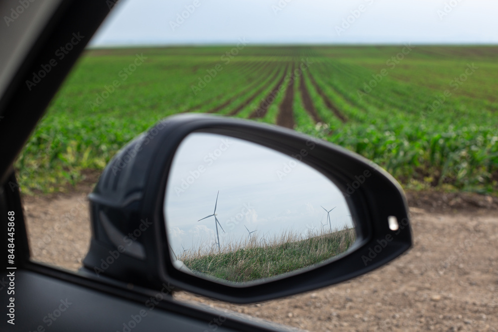 Windmill to produce electricity in cars mirror, behind green plantations grow in the field. Vegetable rows. Farming, agriculture. Landscape with agricultural land. Windmill to produce electricity in cars mirror, behind green plantations grow in the field. Vegetable rows. Farming, agriculture. Landscape with agricultural land.