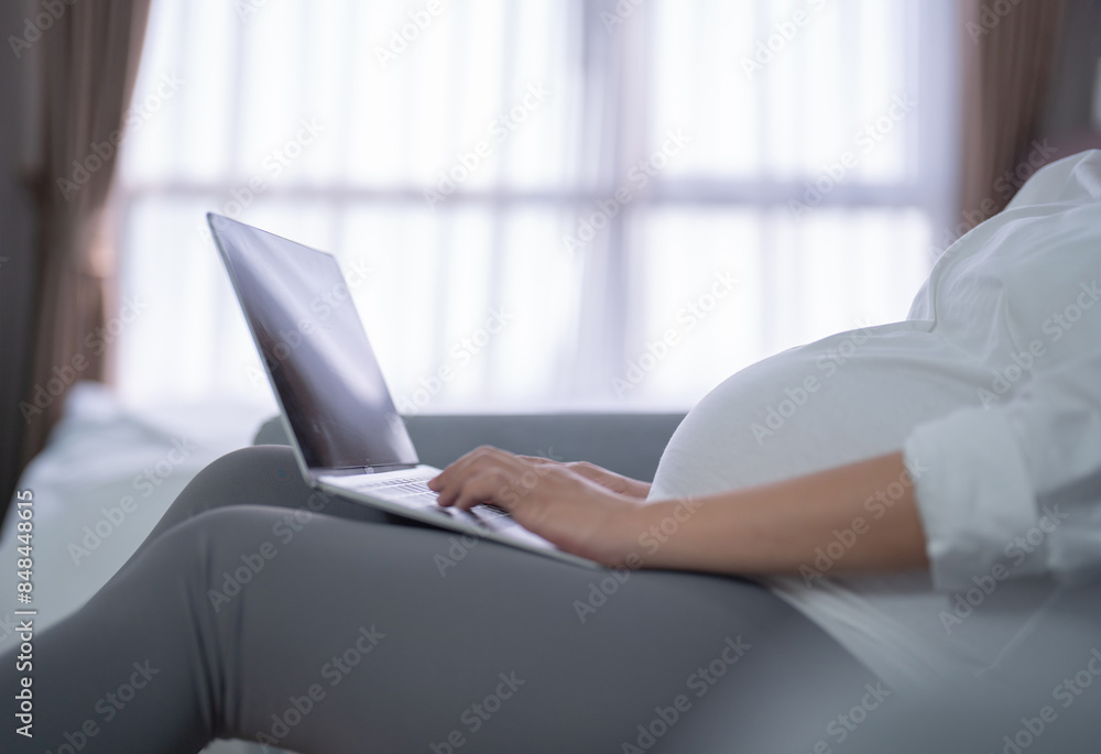 Fototapeta premium A portrait of happy Asian pregnant woman using, working online with computer laptop notebook tablet device with screen in bedroom at home. Having a baby. Family people lifestyle. Mom love.